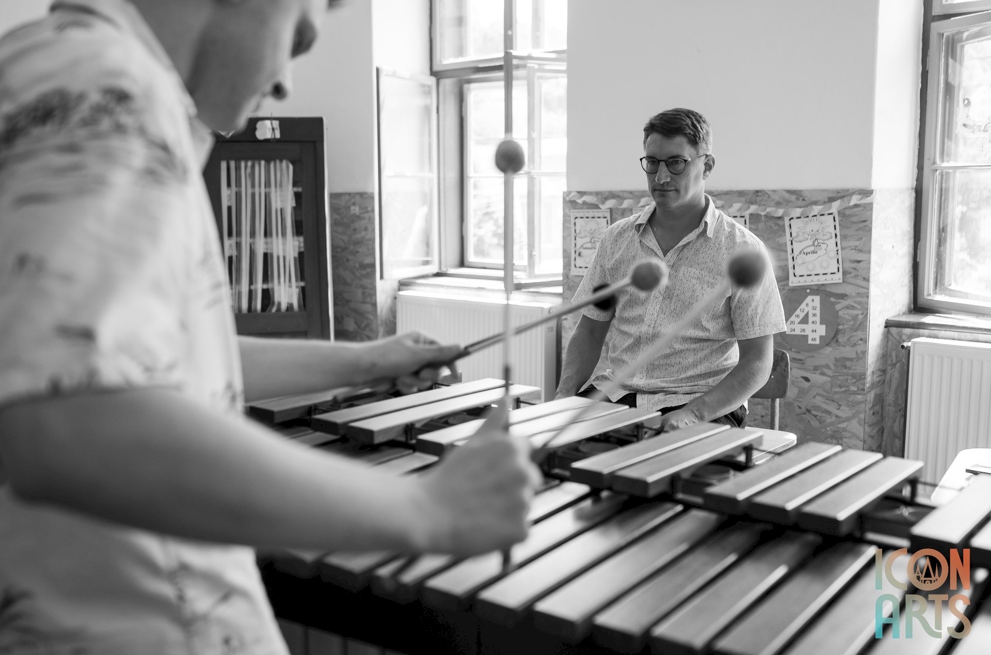 Alexandru Anastasiu ascult&acirc;nd un t&acirc;năr muzician la marimbă, &icirc;ntr-o sală de clasă (alb-negru)